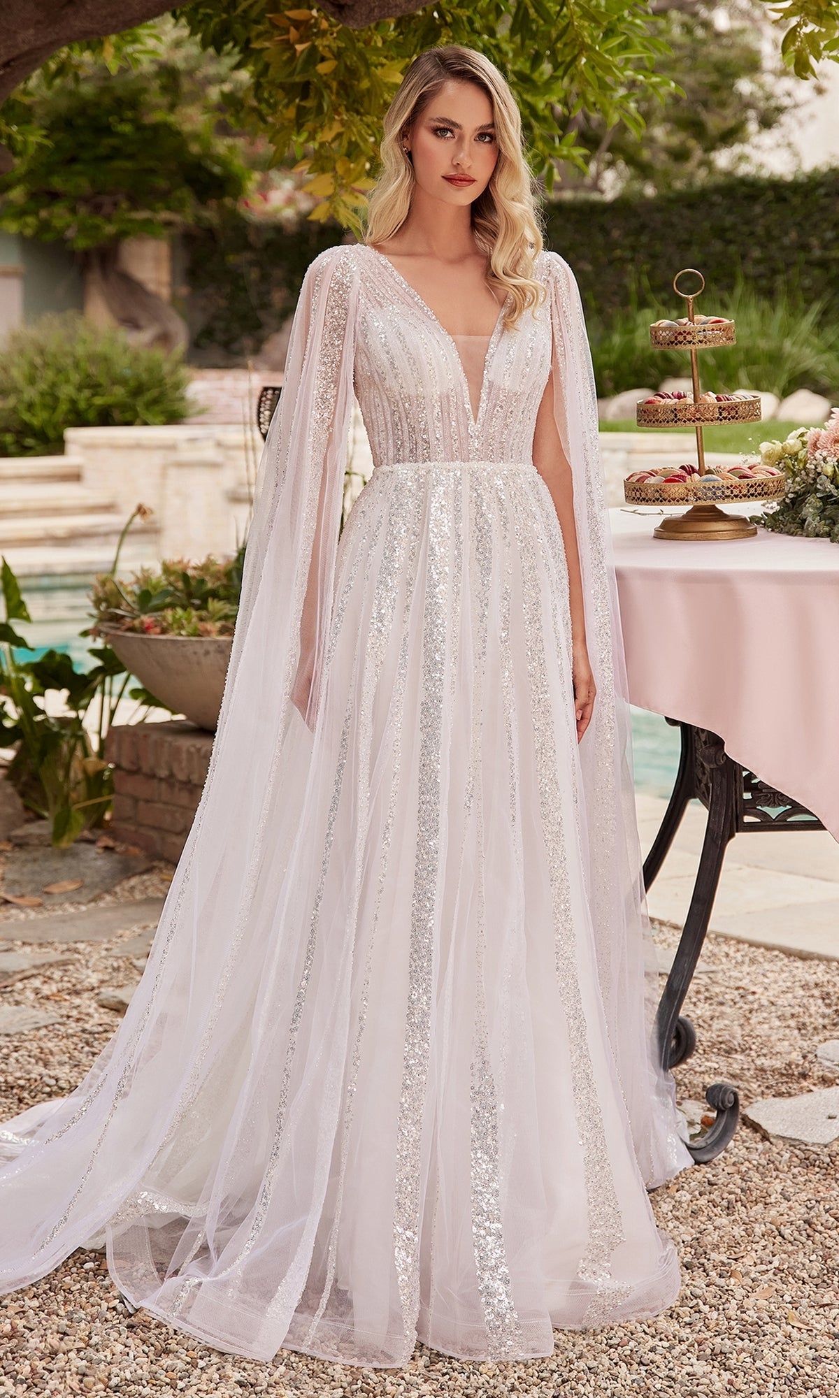 A woman stands outdoors in the Ladivine CD852W Drape-Sleeve White Formal Dress, featuring sheer cape sleeves, next to a table decorated with desserts and flowers.